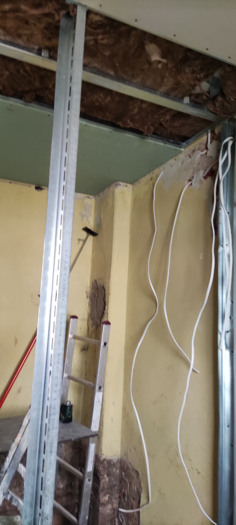 Wnętrze w trakcie remontu: widoczne profile metalowe, ocieplenie poddasza, instalacja elektryczna na żółtej ścianie, drabina aluminiowa i ślady po naprawach na ścianie.