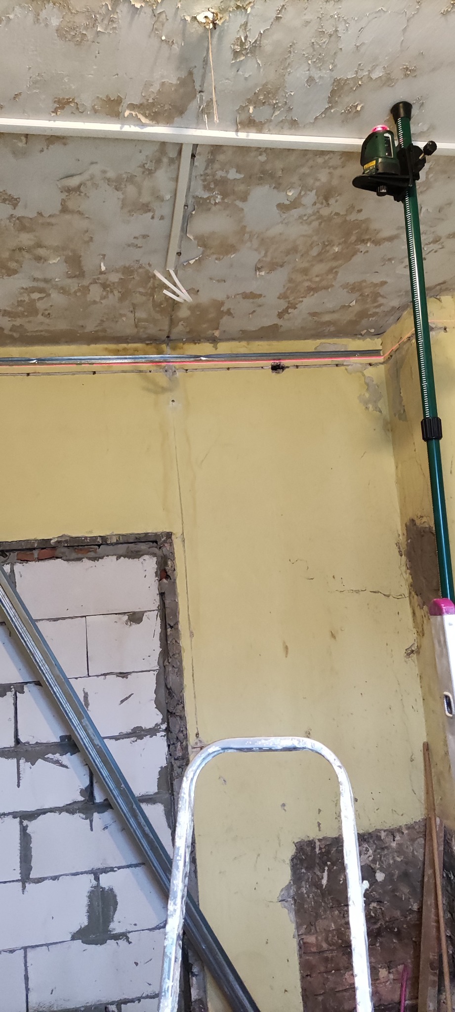 Wnętrze w trakcie prac remontowych: ściana z żółtą farbą, otwór w ścianie z widocznymi bloczkami, drabina, poziomica laserowa wyznaczająca linię na ścianie, sufit z odpadającą farbą.