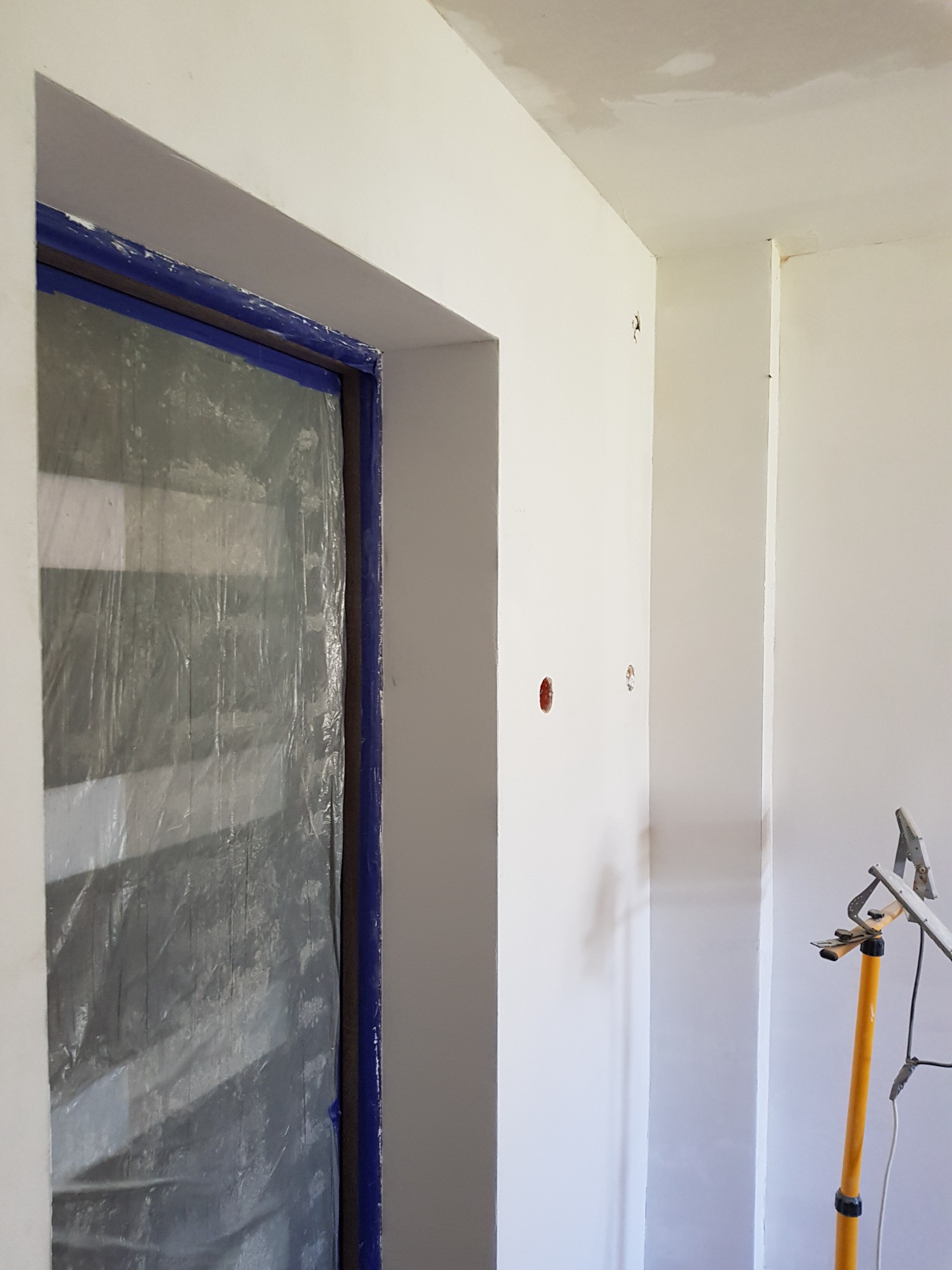 Przygotowane do malowania wnętrze: drzwi zabezpieczone folią i taśmą malarską, ściany po szpachlowaniu, widoczne punkty elektryczne, żółty statyw oświetleniowy.