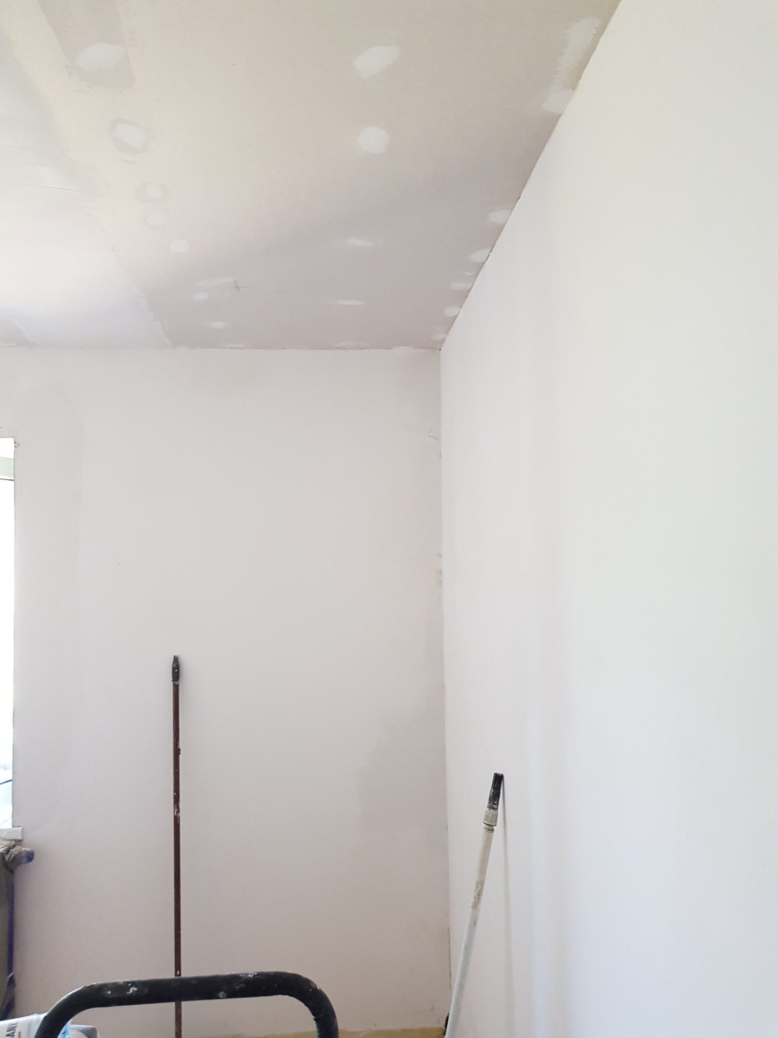 Świeżo pomalowane wnętrze z widocznymi łatami szpachli na suficie, przygotowane do dalszych prac wykończeniowych, widoczne narzędzia malarskie.