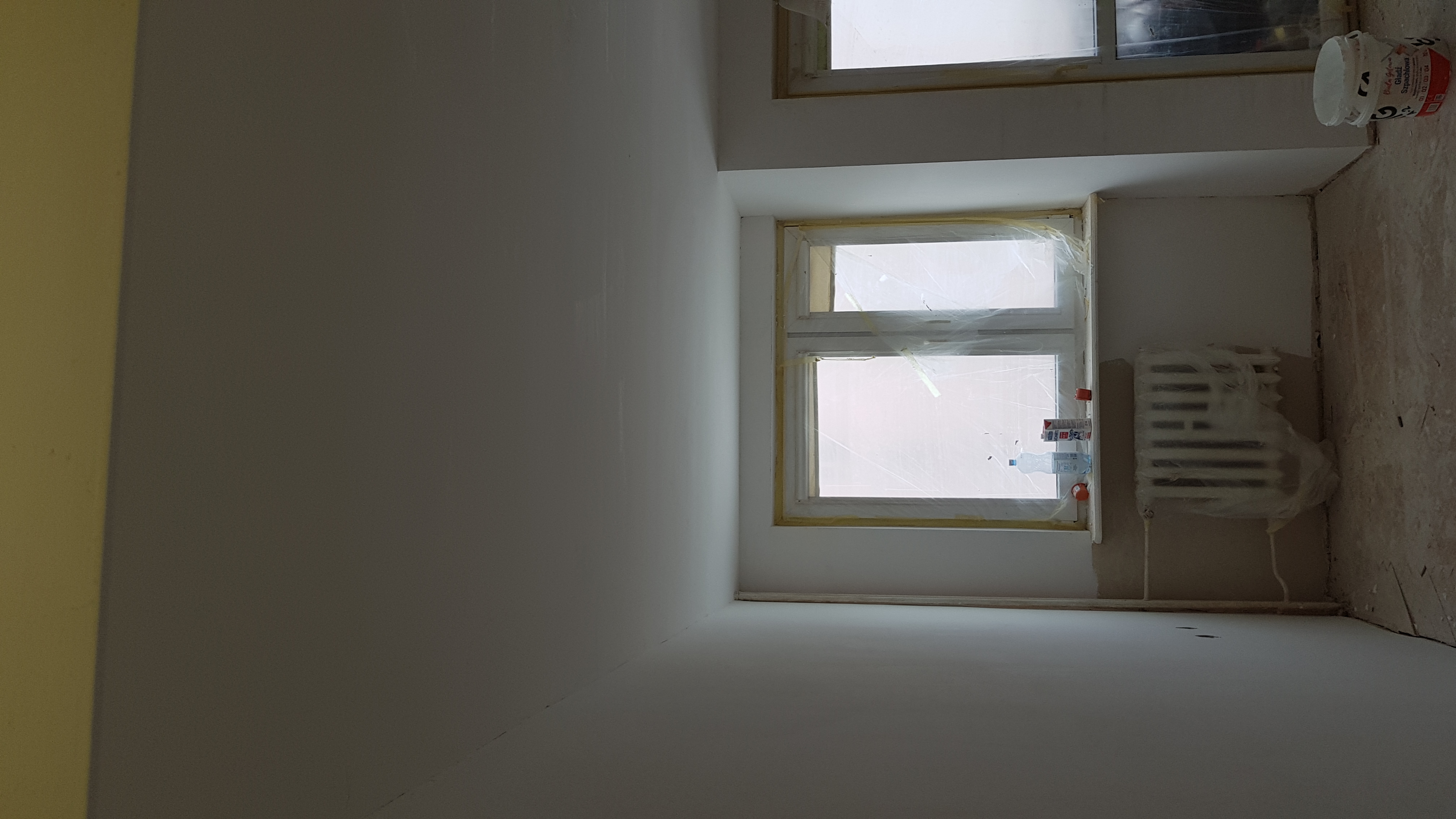 Wnętrze pokoju w trakcie remontu: świeżo pomalowane ściany na biało, okna i grzejnik zabezpieczone folią ochronną, widoczne ślady po szpachlowaniu i wiadro z resztkami gładzi.