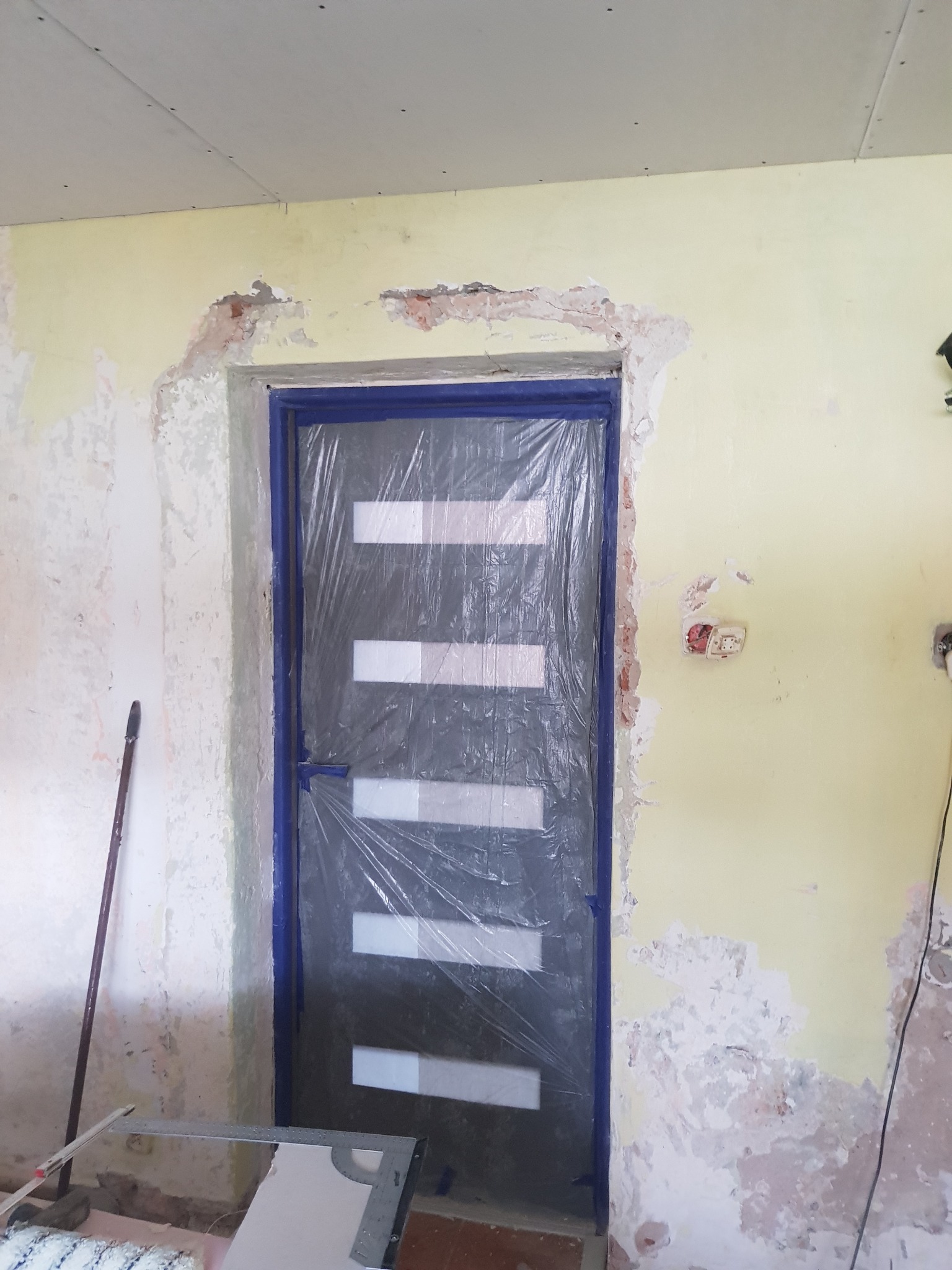 Przygotowanie do wykończenia ściany wewnątrz pomieszczenia: zabezpieczone folią drzwi, ściana z ubytkami i pozostałościami starej farby, częściowo zagruntowana płyta gipsowa na suficie, widoczny...