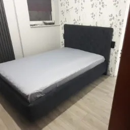 Ciemnoszare łóżko tapicerowane z pikowanym zagłówkiem, ustawione w pokoju z jasnymi panelami podłogowymi i wzorzystą tapetą w drobne listki.