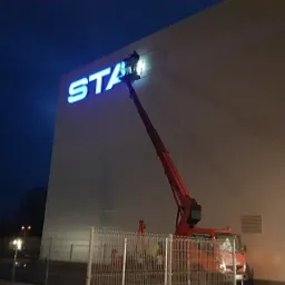 Montaż podświetlanego logo 'STA' na elewacji budynku przemysłowego z użyciem podnośnika koszowego o zmierzchu.