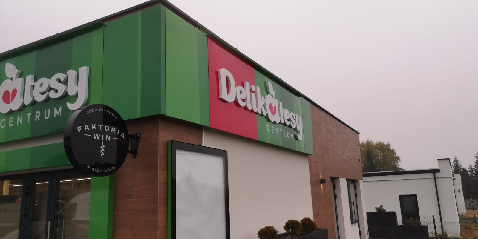 Zewnętrzna identyfikacja wizualna sklepu Delikatesy Centrum z podświetlanym szyldem Faktoria Win po lewej stronie.