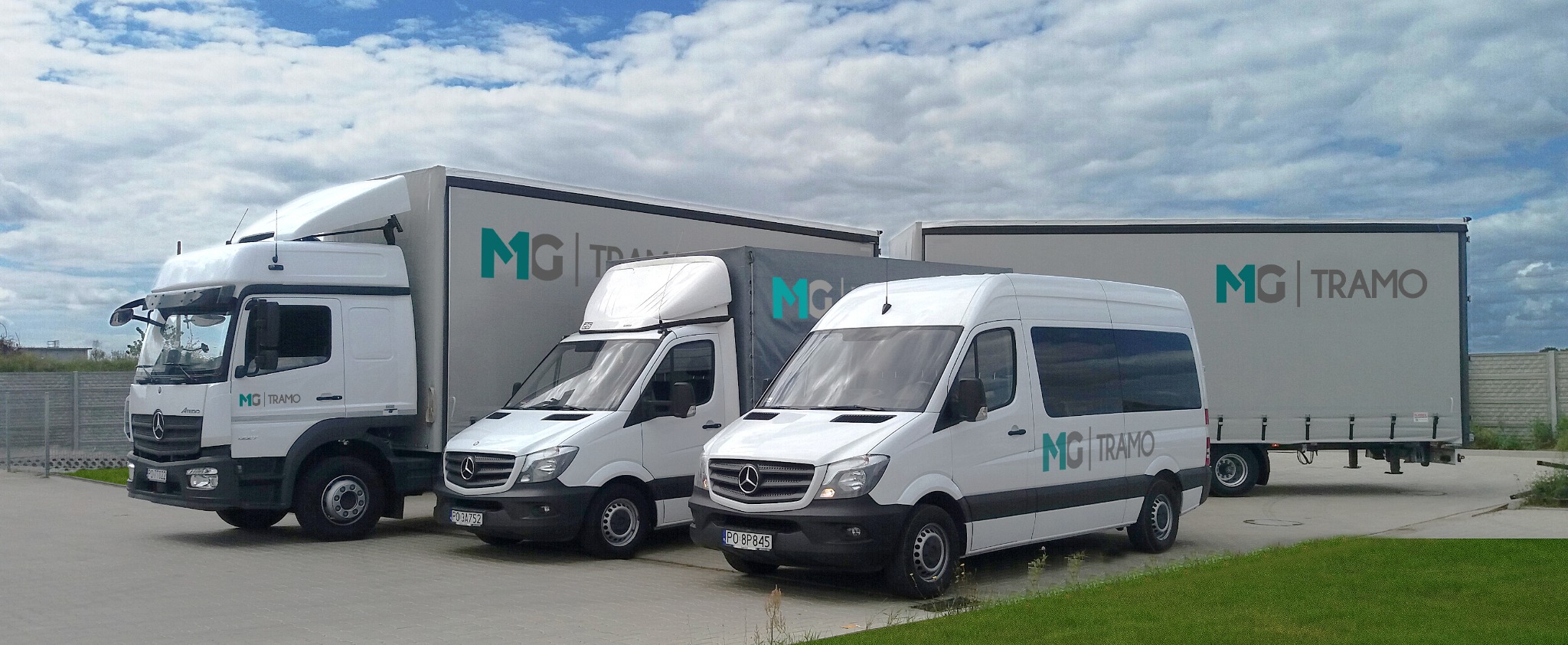 Flota pojazdów firmy MG Tramo: ciągnik siodłowy, bus i furgonetka z logo firmy na tle pochmurnego nieba.