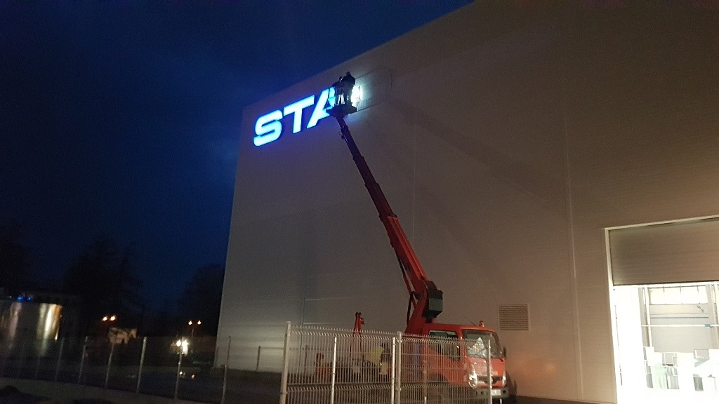 Montaż podświetlanego logo 'STA' na elewacji budynku przemysłowego z użyciem podnośnika koszowego o zmierzchu.