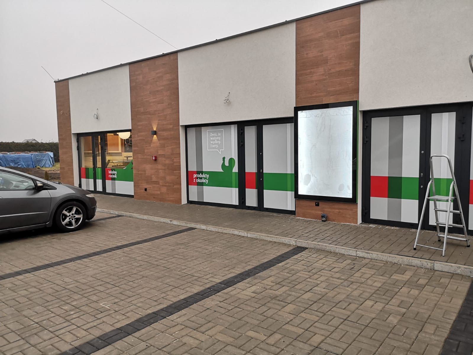 Fasada sklepu spożywczego z grafiką na oknach i drabiną opartą o ścianę, widok z parkingu w pochmurny dzień.