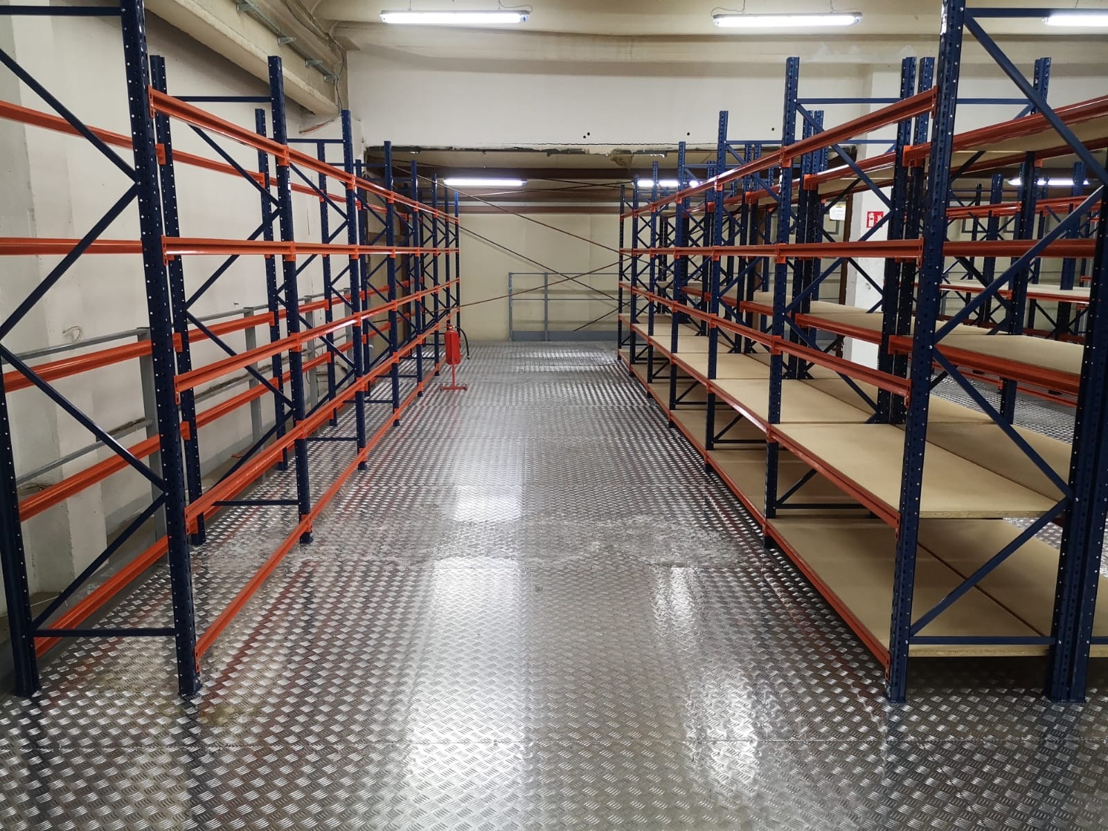 Puste, metalowe regały magazynowe z pomarańczowymi poprzeczkami i niebieskimi słupkami na metalowej podłodze z fakturą rombów, oświetlone jarzeniówkami.