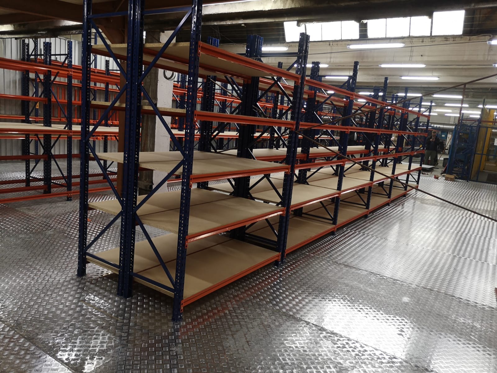 Metalowe regały magazynowe z niebieskimi słupkami i pomarańczowymi poprzeczkami, z drewnianymi półkami, ustawione w hali z metalową podłogą o wzorze rombów.
