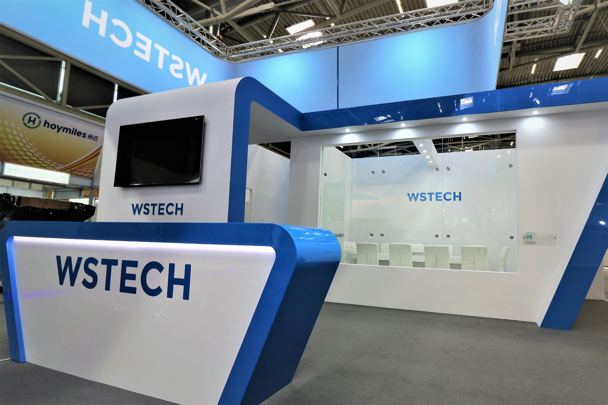 Nowoczesne stoisko targowe WSTECH w kolorach białym i niebieskim, z podświetlanym logo, ekranem i przeszkloną salą konferencyjną w tle.