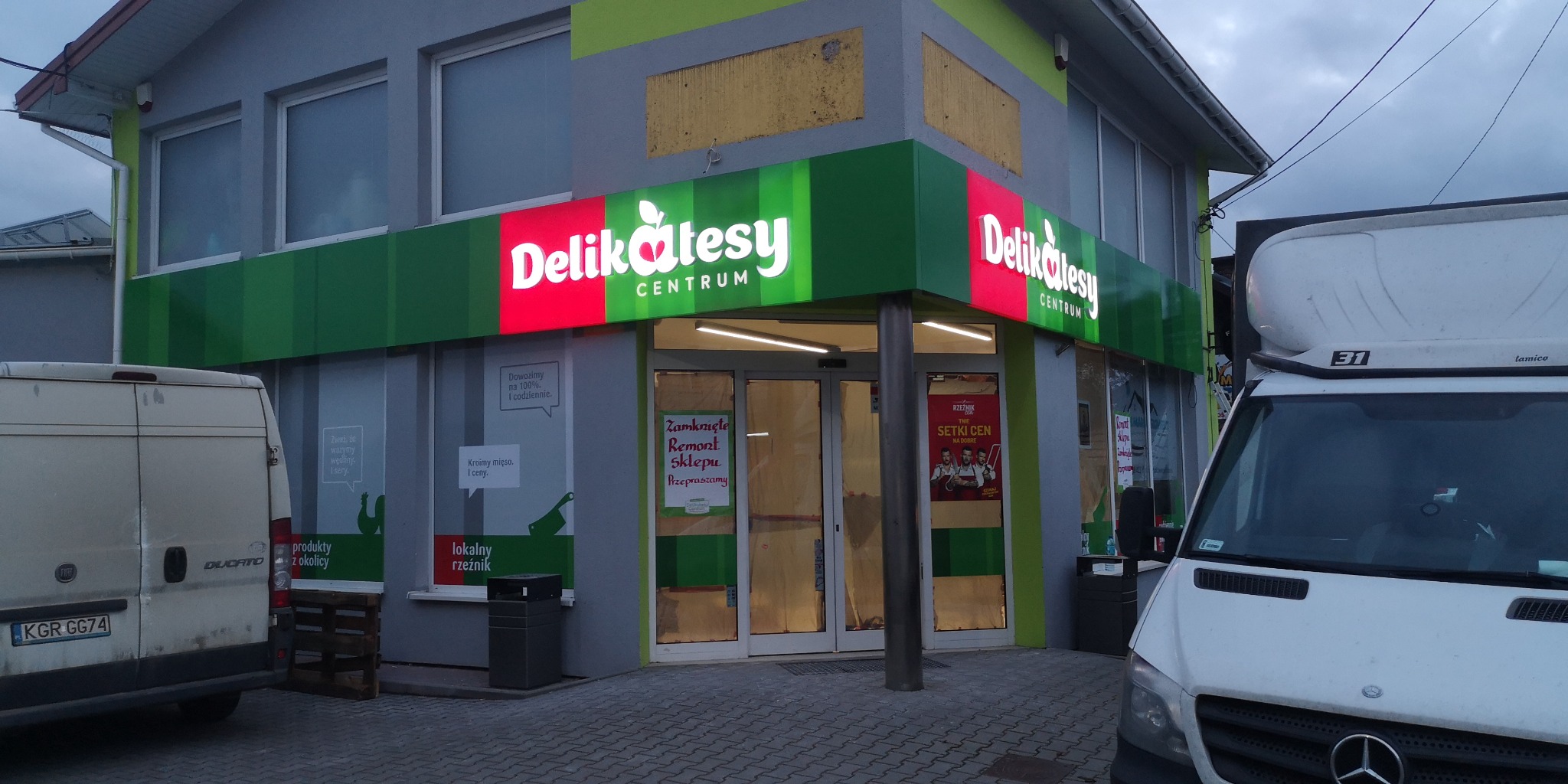 Oświetlony szyld sklepu spożywczego 'Delikatesy Centrum' z zielono-czerwonym tłem i białym napisem, widoczny z zewnątrz, podczas remontu, z zaparkowanymi samochodami dostawczymi po bokach.