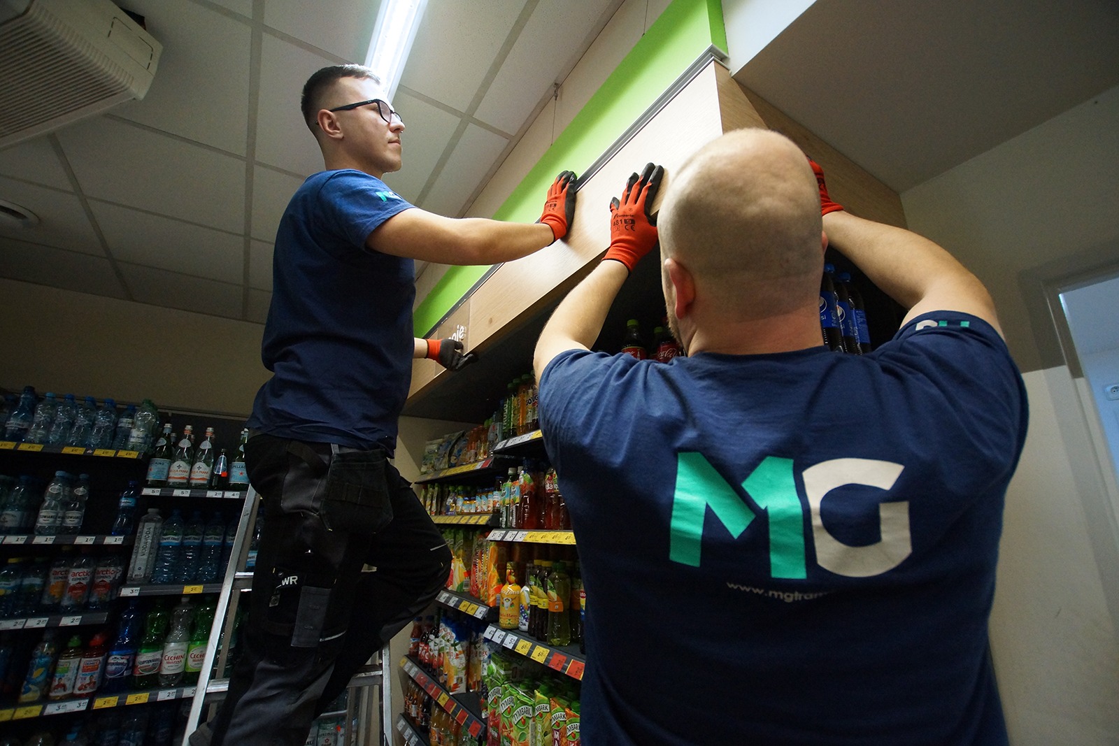 Dwóch mężczyzn w pomarańczowych rękawicach montuje drewnianą zabudowę nad półkami sklepowymi, jeden stoi na drabinie, w tle widoczne produkty spożywcze.