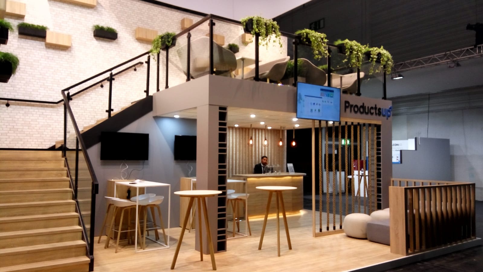 Aranżacja stoiska targowego z drewnianymi schodami, roślinami doniczkowymi na ścianie, barem z oświetleniem i ekranem z napisem ProductsUp.