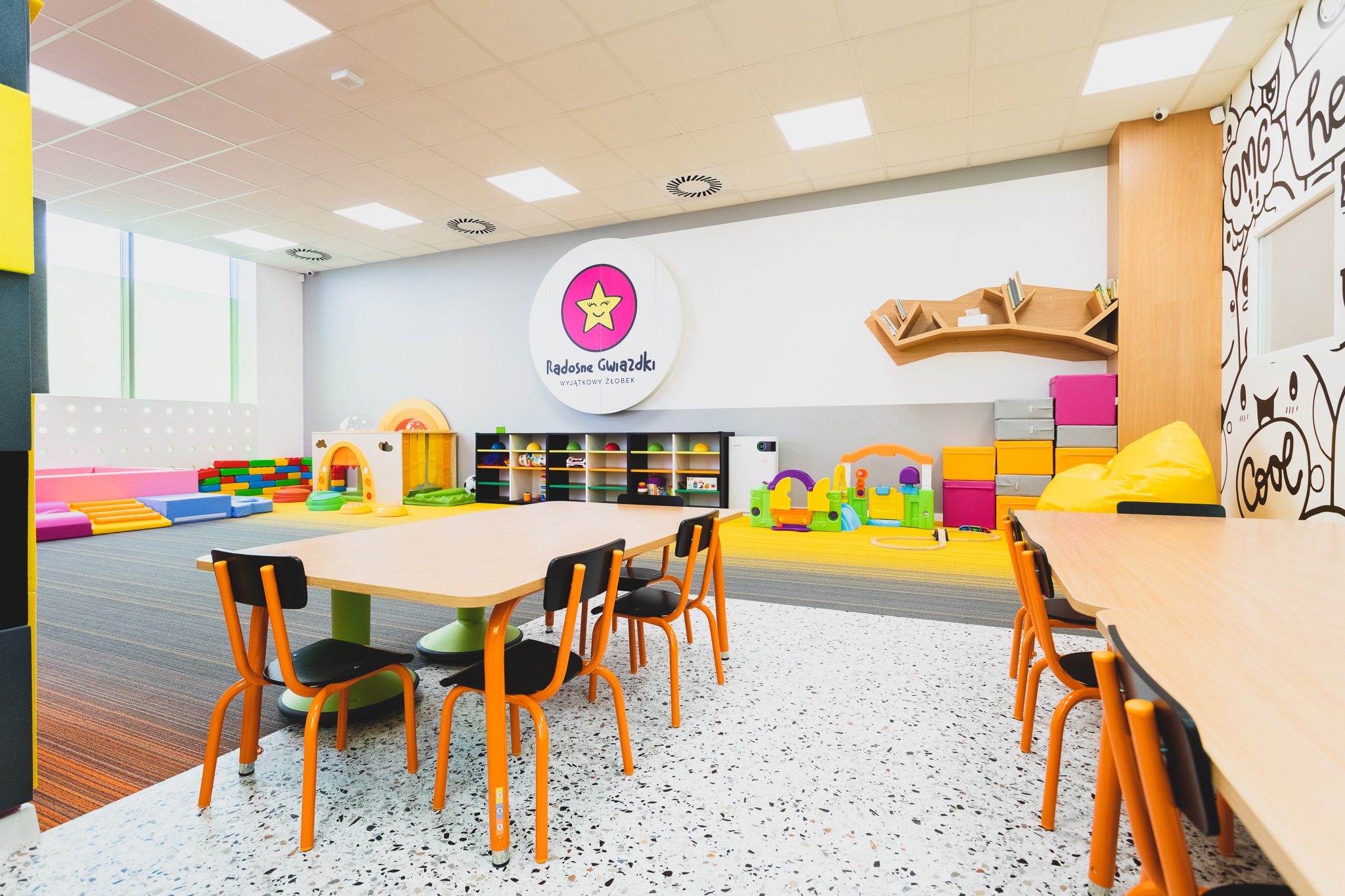 Jasne, przestronne wnętrze sali zabaw z kolorowymi meblami, stolikami z pomarańczowymi krzesłami, regałami z zabawkami i miękkimi elementami do zabawy, logo placówki na ścianie.