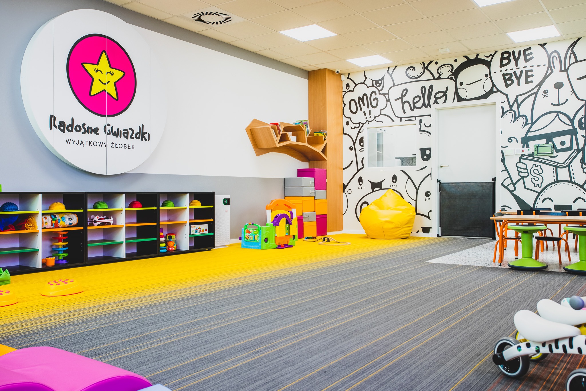 Jasne wnętrze sali zabaw z kolorowymi zabawkami, regałami, pufą i ścianą z rysunkami, widoczny fragment logo firmy.