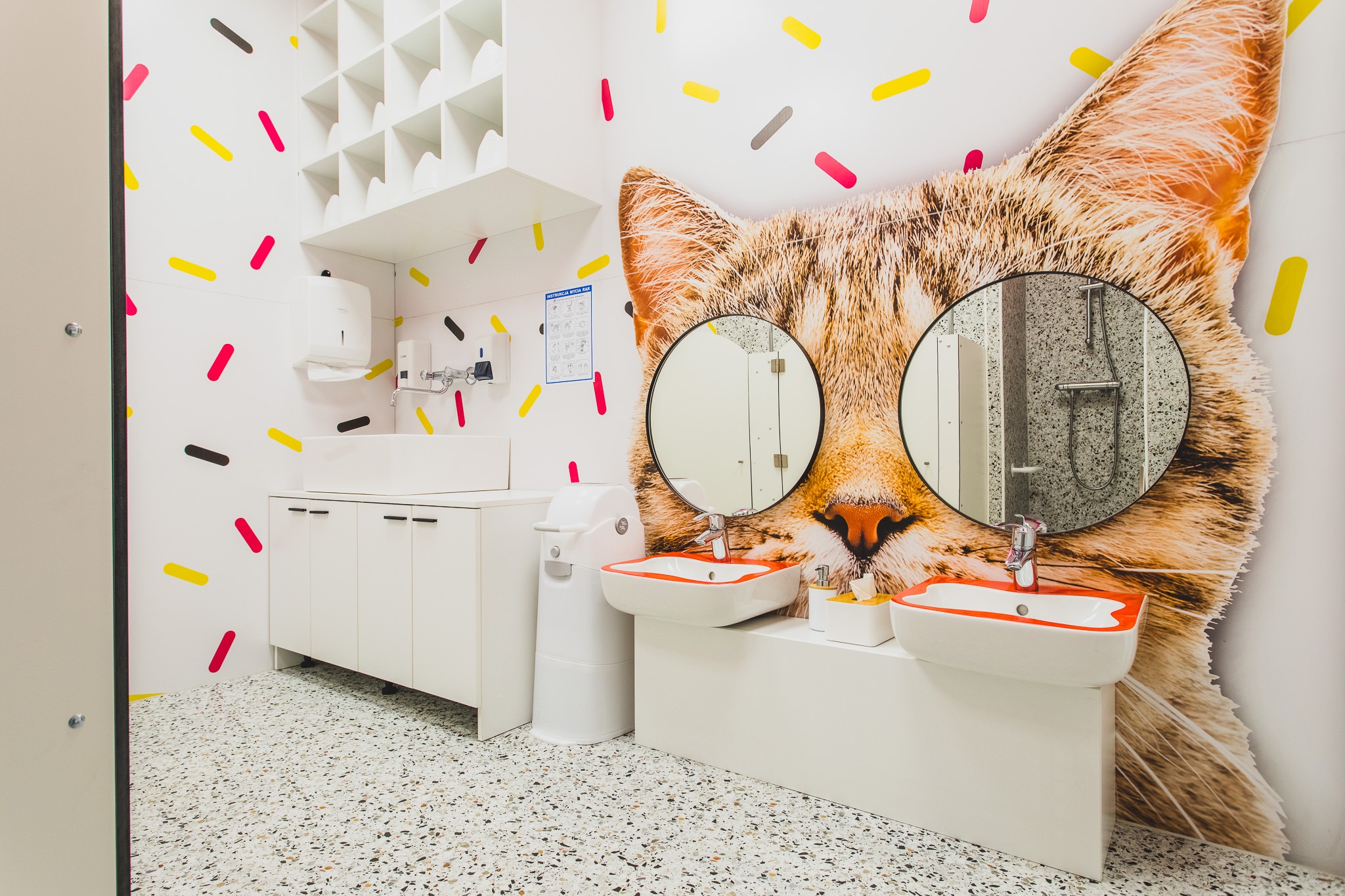 Umywalki z pomarańczowymi akcentami w łazience dziecięcej z motywem kota na ścianie i wzorem posypki na ścianach i podłodze.