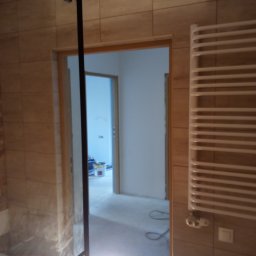 Roberto bud - Częściowo wykończona łazienka z beżowymi płytkami, widoczna kabina prysznicowa z czarnymi profilami i grzejnik drabinkowy. W tle otwarte drzwi do innego pomieszczenia z wiadrem farby.