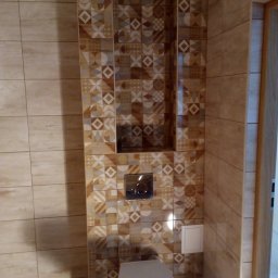 Roberto bud - Wykończona toaleta z mozaiką z płytek w odcieniach beżu i brązu, wnęka w ścianie nad spłuczką, widoczny fragment drzwi.