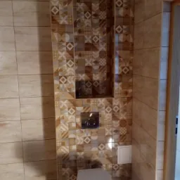 Wykończona toaleta z mozaiką z płytek w odcieniach beżu i brązu, wnęka w ścianie nad spłuczką, widoczny fragment drzwi.