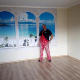 Roberto bud - Mężczyzna w ubrudzonym farbą czerwonym kombinezonie stoi na tle ściany z fototapetą przedstawiającą widok na morze z kolumnami, w pustym pokoju z panelami na podłodze.