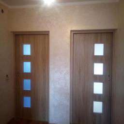 Roberto bud - Dwa zamontowane, drewnopodobne drzwi wewnętrzne z trzema kwadratowymi przeszkleniami, jedno z przeszkleniami w kolorze niebieskim, drugie z białymi, na tle ściany z efektem dekoracyjnym, oświetlone...