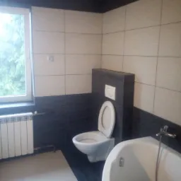 Wykończona łazienka z biało-czarnymi płytkami, widoczna toaleta z zabudowanym systemem spłukiwania, wanna narożna i grzejnik pod oknem, z widokiem na drzewa za oknem.