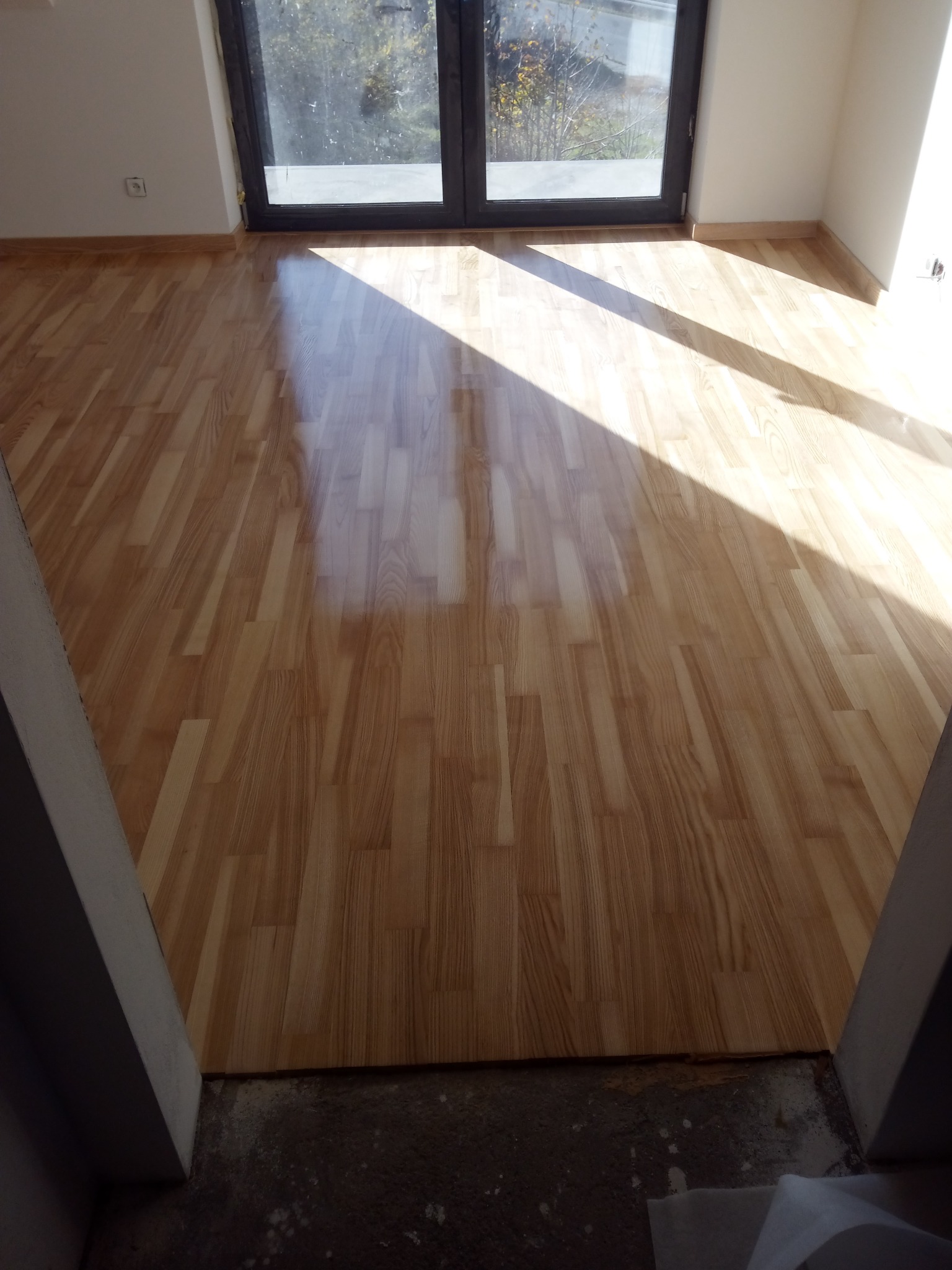 Świeżo położona podłoga z paneli laminowanych w jasnym odcieniu drewna, widoczna przez framugę drzwi, z silnym światłem słonecznym padającym z okna balkonowego.