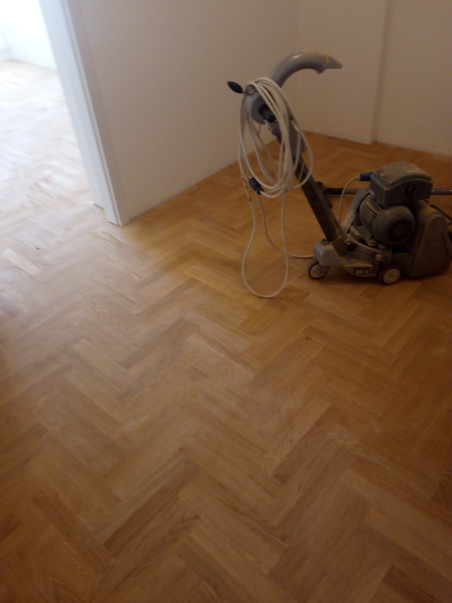 Parkiet ułożony w jodełkę po renowacji, widoczna szlifierka do parkietu z przewodem, jasne drewno, neutralne tło ścian.