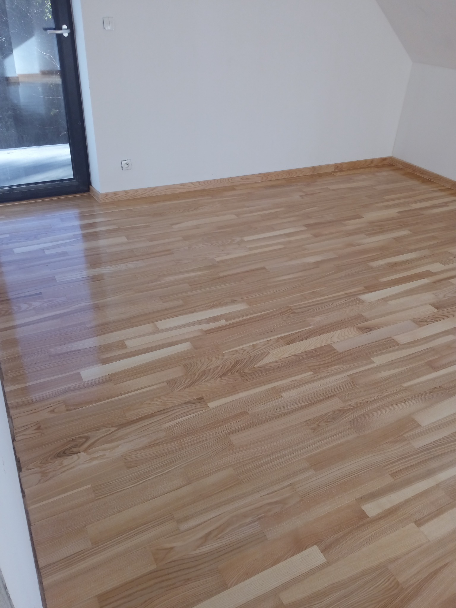 Jasna, nowa podłoga parkietowa ułożona w jodełkę w pustym pokoju z białymi ścianami i czarną ramą okienną.