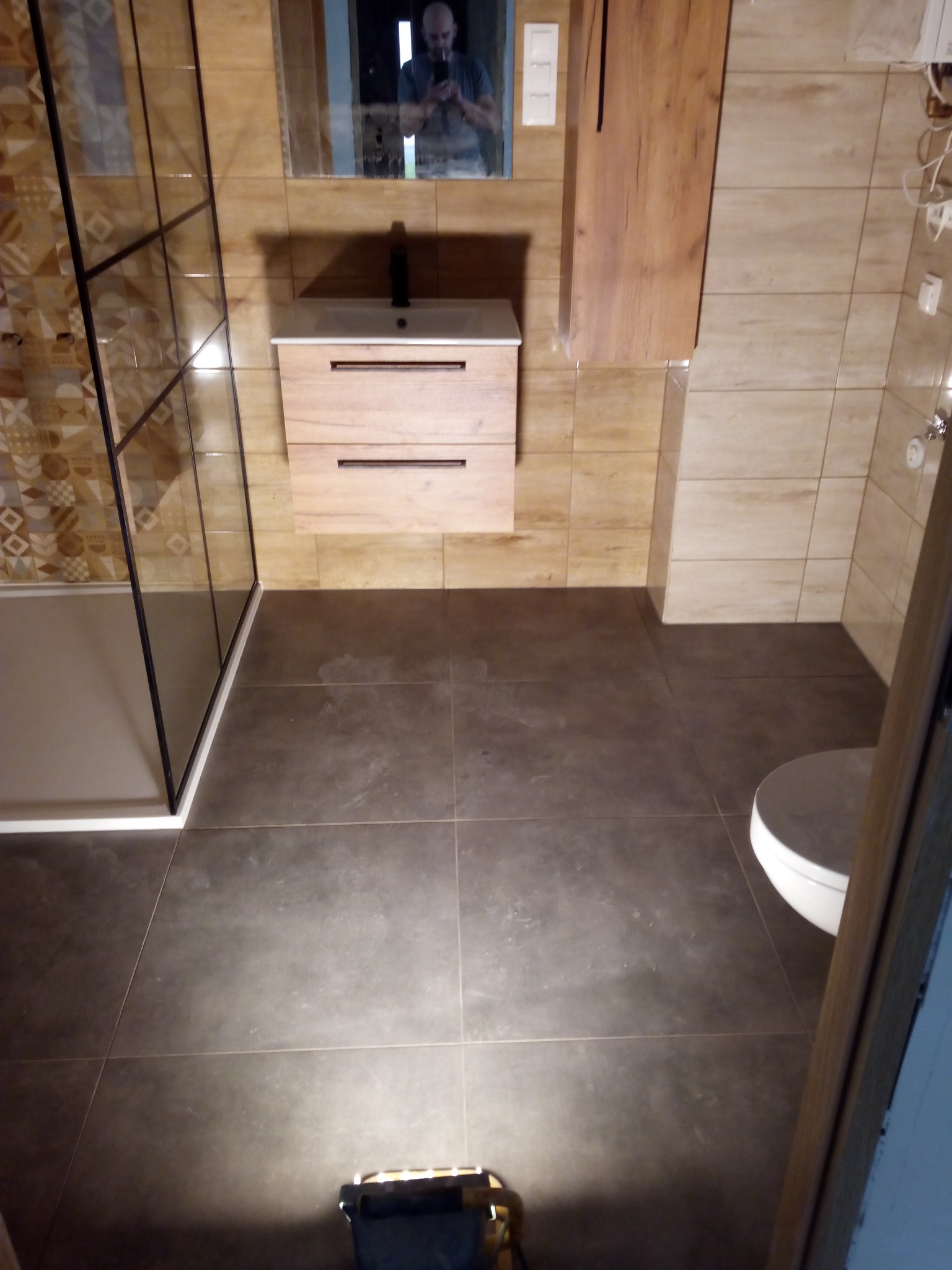 Nowoczesna łazienka z kabiną prysznicową typu walk-in z czarnymi profilami, podwieszaną szafką z umywalką o drewnianym froncie, brązowymi płytkami na ścianach i podłodze oraz toaletą w rogu, widok...