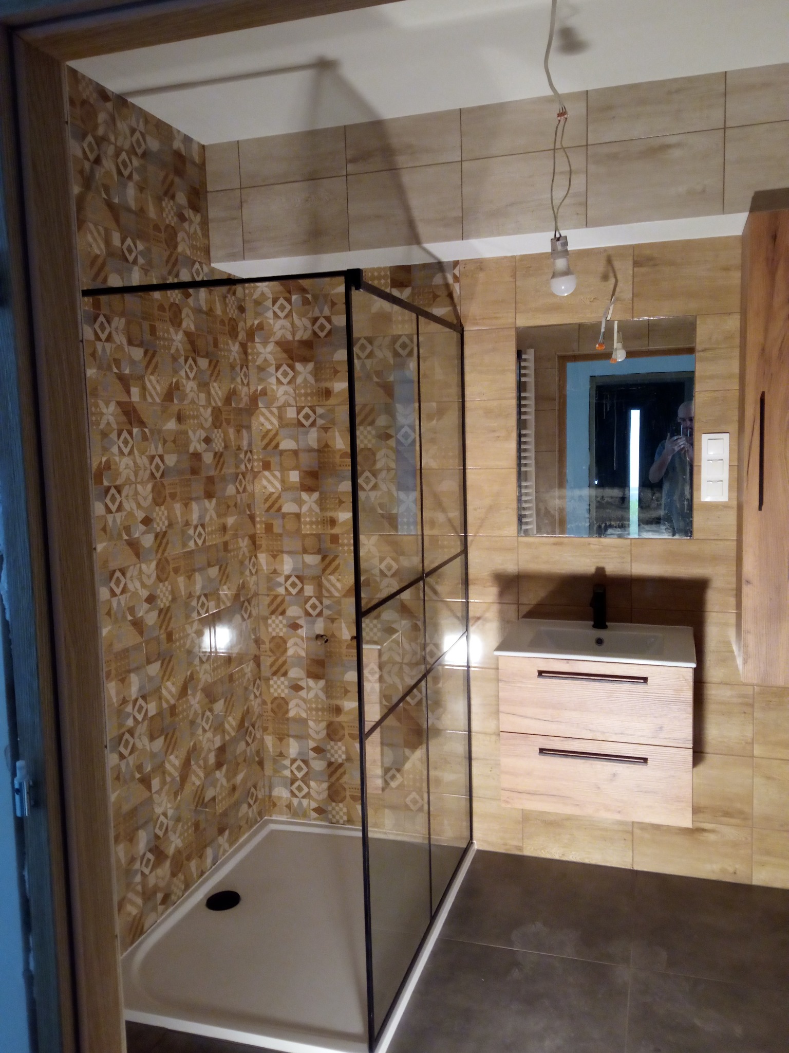 Nowoczesna kabina prysznicowa z czarnymi profilami, wzorzyste płytki ścienne w odcieniach brązu, umywalka z drewnopodobną szafką, lustro z odbiciem osoby robiącej zdjęcie, sufit z niedokończonym...