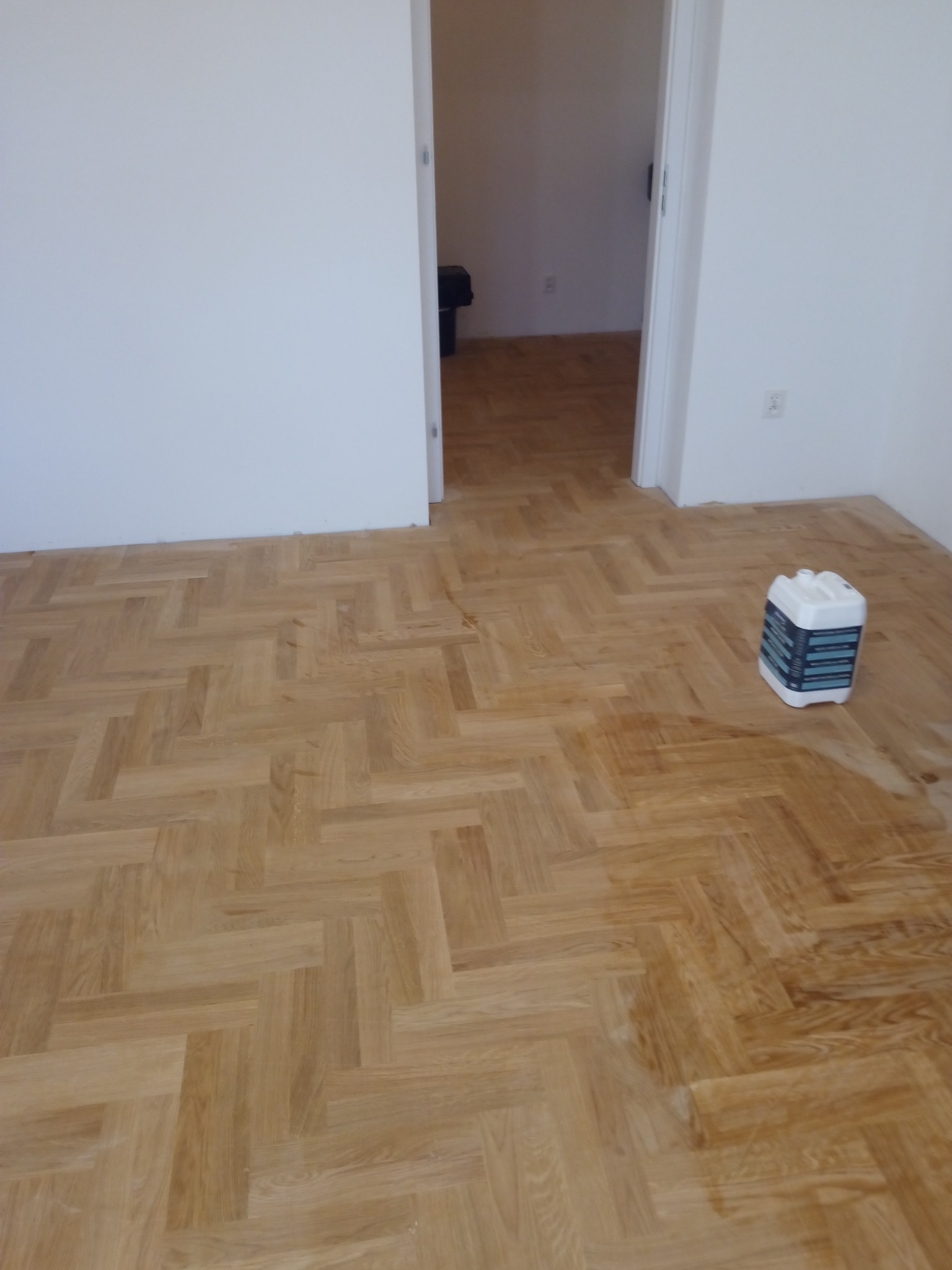 Parkiet ułożony w jodełkę, częściowo pokryty lakierem lub olejem, z widocznym pojemnikiem z preparatem do konserwacji drewna w rogu pokoju.