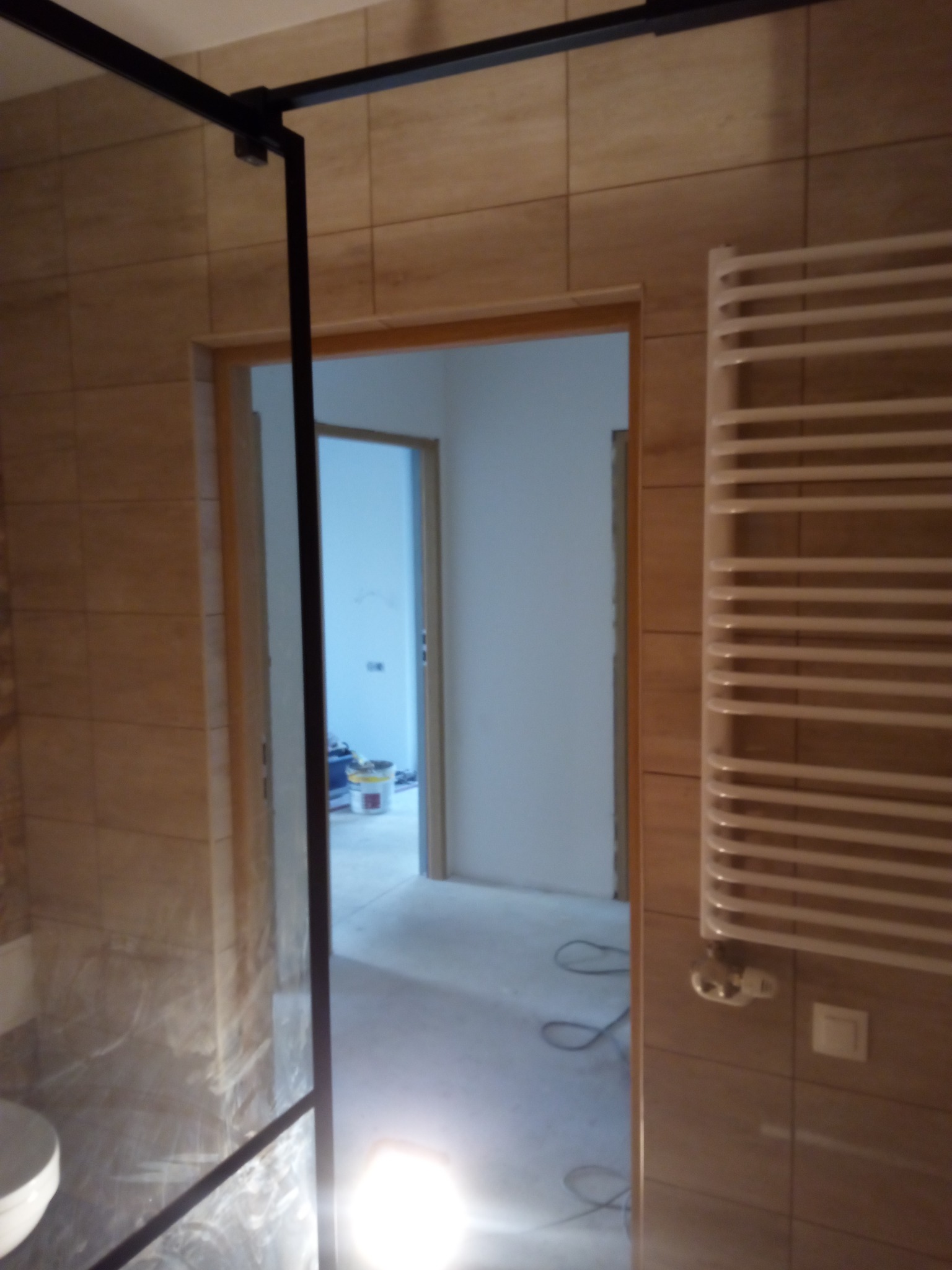 Częściowo wykończona łazienka z beżowymi płytkami, widoczna kabina prysznicowa z czarnymi profilami i grzejnik drabinkowy. W tle otwarte drzwi do innego pomieszczenia z wiadrem farby.