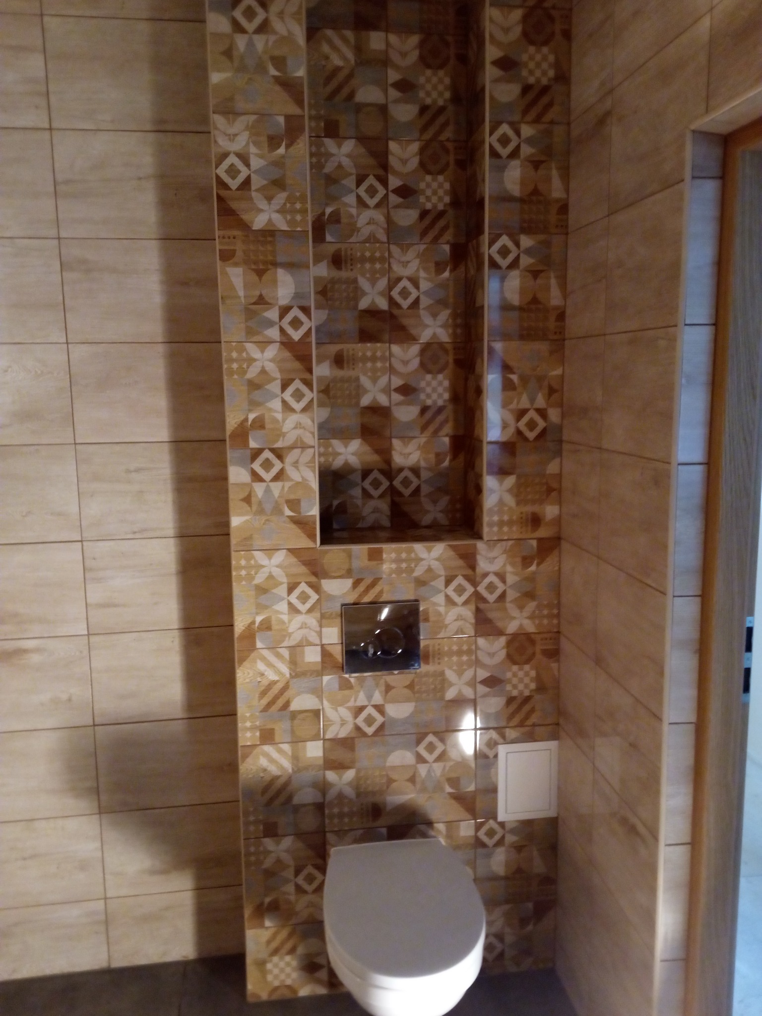 Wykończona toaleta z mozaiką z płytek w odcieniach beżu i brązu, wnęka w ścianie nad spłuczką, widoczny fragment drzwi.