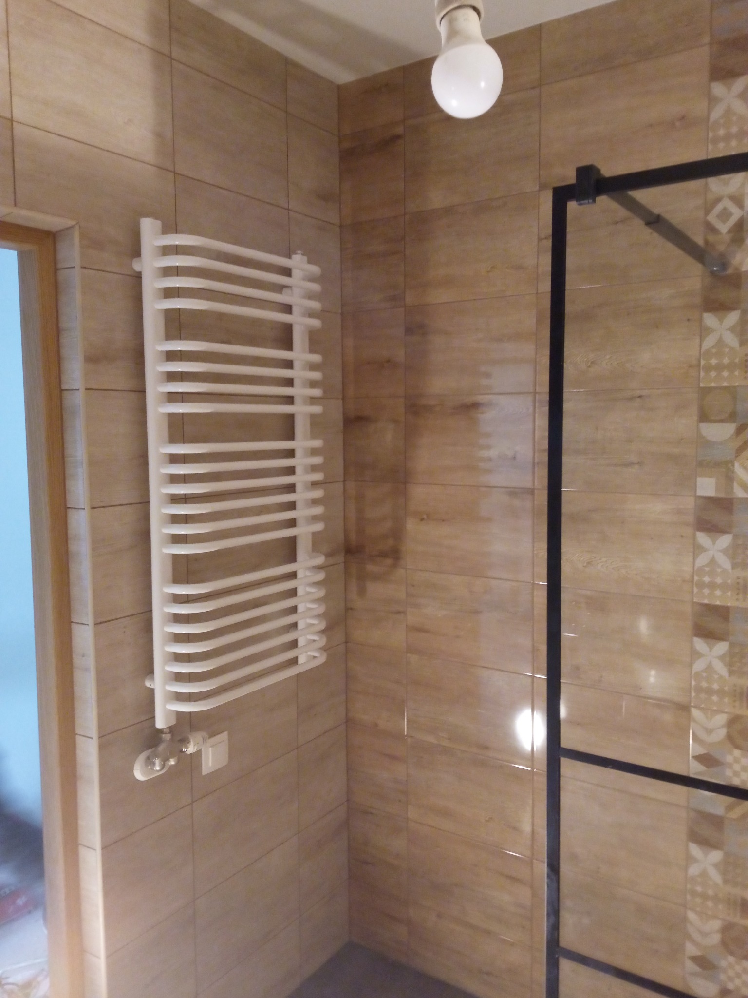 Wykończona łazienka z beżowymi płytkami imitującymi drewno, biały grzejnik drabinkowy i czarna rama kabiny prysznicowej z ozdobnymi panelami.