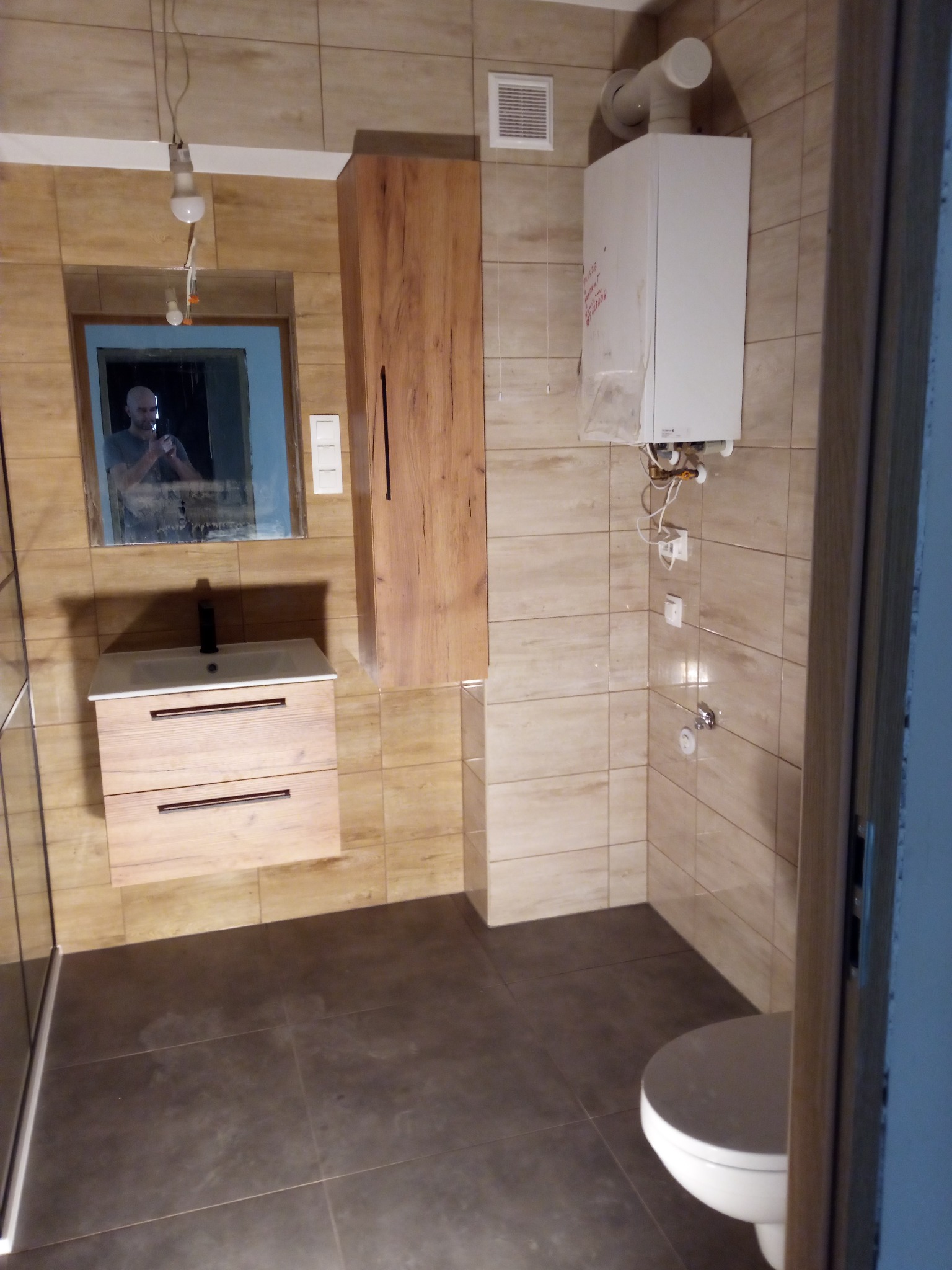 Łazienka z beżowymi płytkami, wiszącą szafką z umywalką z drewnianymi frontami, białym bojlerem i lustrem, w którym odbija się osoba robiąca zdjęcie.