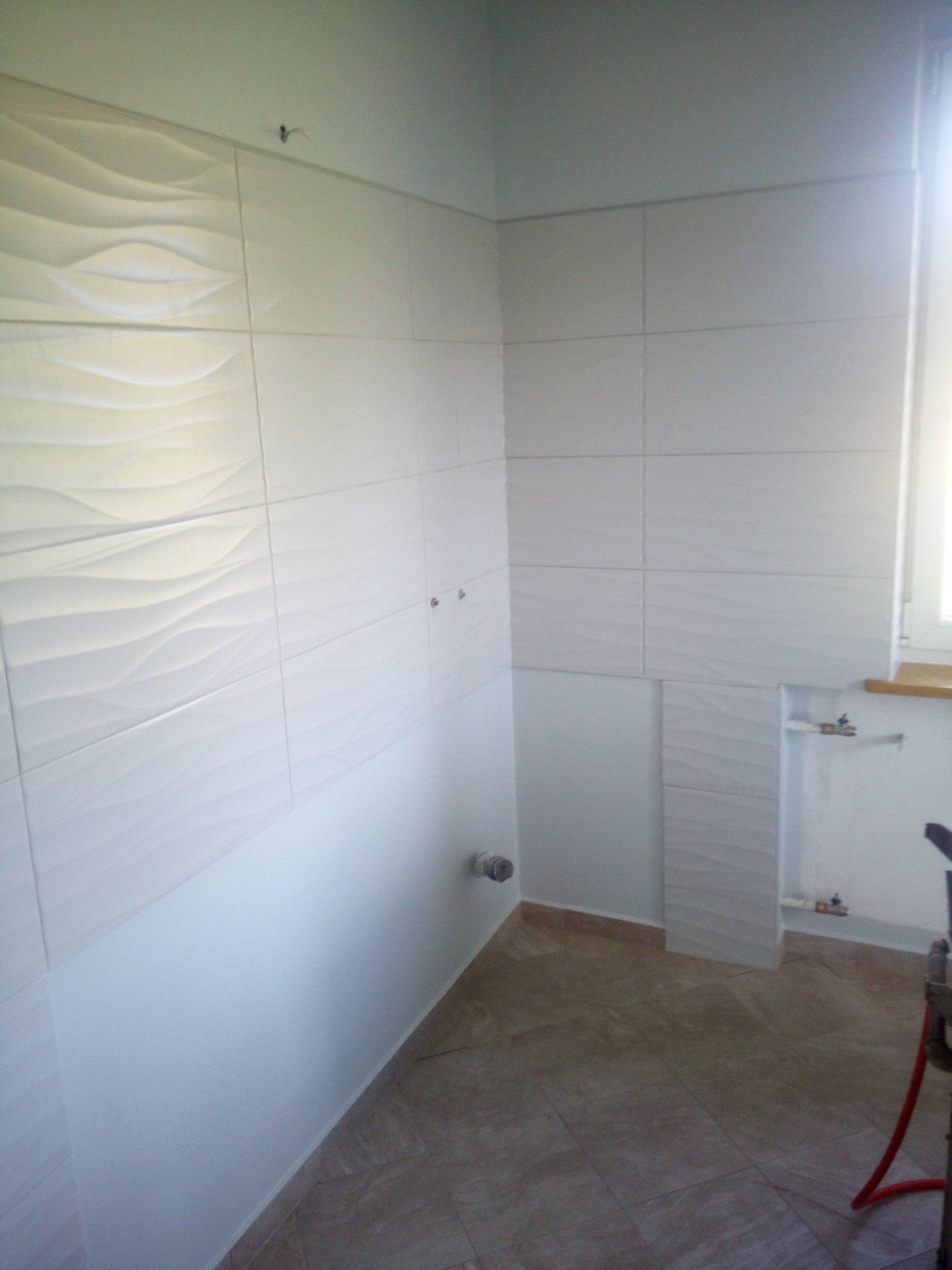 Wnętrze łazienki w trakcie remontu, widoczne białe płytki ścienne z falistym wzorem, brązowe płytki podłogowe, wystające rury i element instalacji, surowy stan wykończenia.
