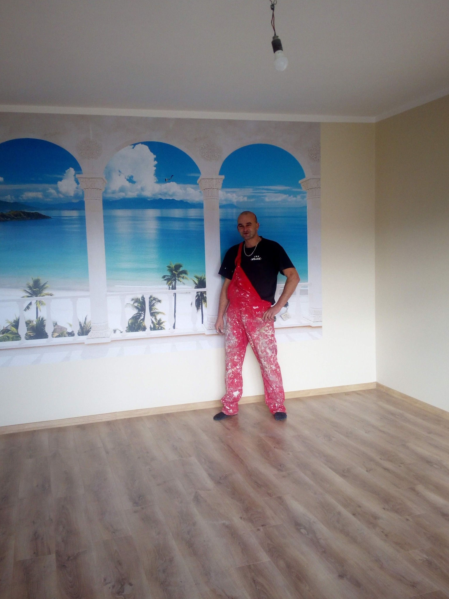Mężczyzna w ubrudzonym farbą czerwonym kombinezonie stoi na tle ściany z fototapetą przedstawiającą widok na morze z kolumnami, w pustym pokoju z panelami na podłodze.
