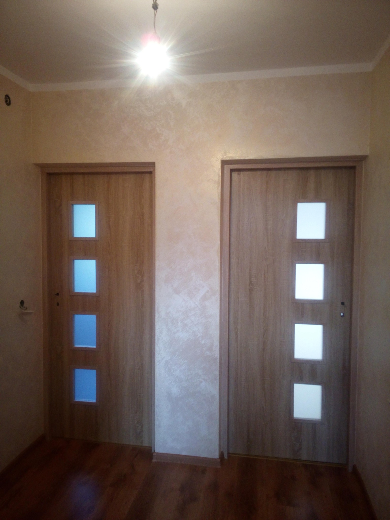 Dwa zamontowane, drewnopodobne drzwi wewnętrzne z trzema kwadratowymi przeszkleniami, jedno z przeszkleniami w kolorze niebieskim, drugie z białymi, na tle ściany z efektem dekoracyjnym, oświetlone...