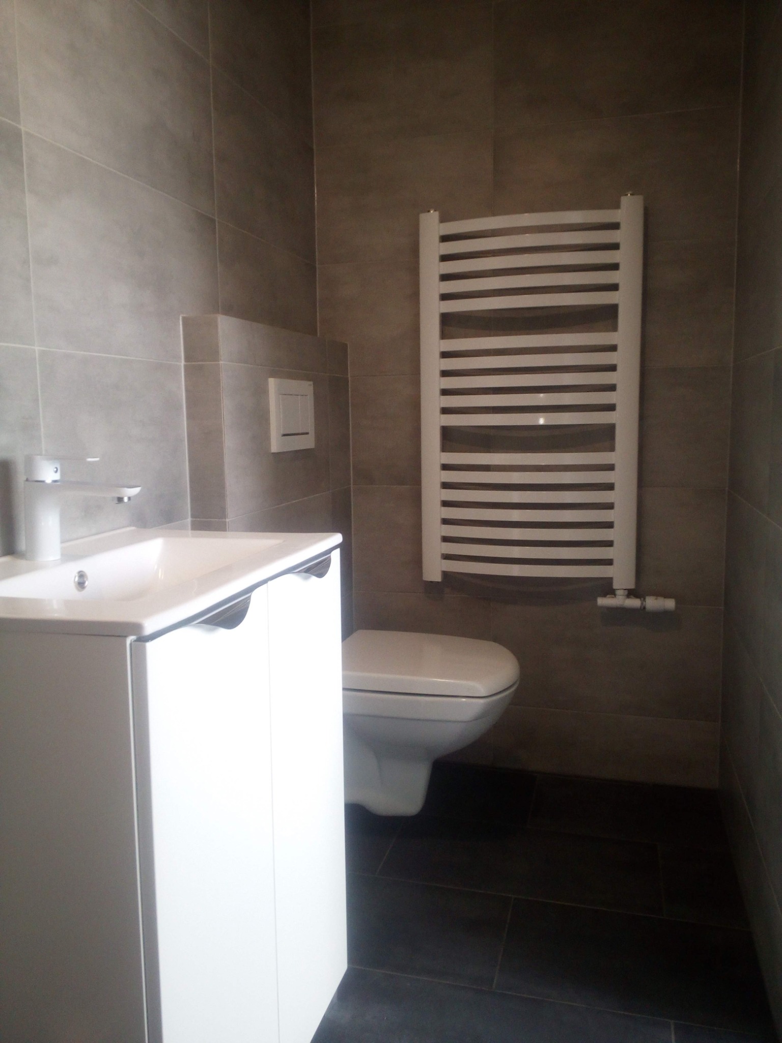 Wykończona łazienka z szarymi płytkami, białą umywalką z szafką, toaletą i grzejnikiem drabinkowym.