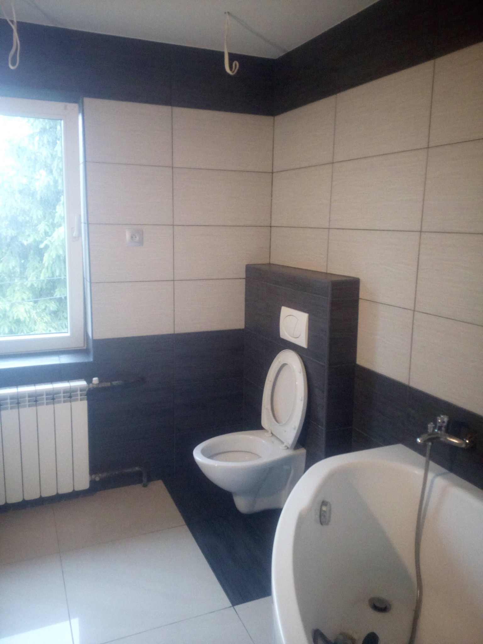 Wykończona łazienka z biało-czarnymi płytkami, widoczna toaleta z zabudowanym systemem spłukiwania, wanna narożna i grzejnik pod oknem, z widokiem na drzewa za oknem.