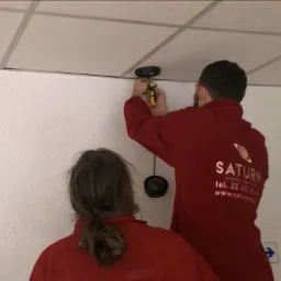 Dwóch instalatorów w czerwonych bluzach z logo firmy montuje czujnik na suficie podwieszanym za pomocą śrubokręta.