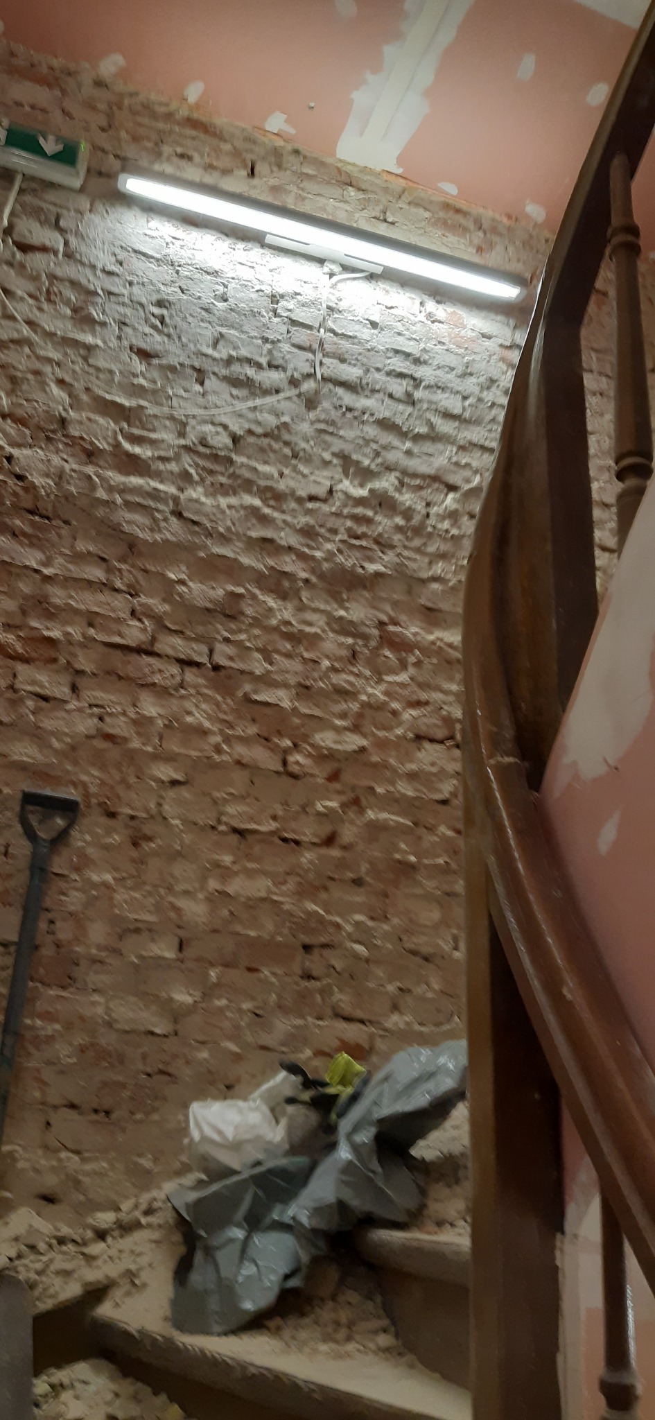 Ceglana ściana w trakcie remontu, widoczna łopata, gruz w workach, oświetlenie jarzeniowe i fragment drewnianej balustrady schodów.