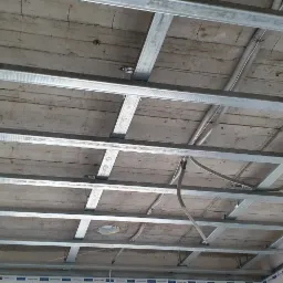 Metalowa konstrukcja podwieszanego sufitu zamontowana pod drewnianym stropem, widoczne przewody elektryczne.