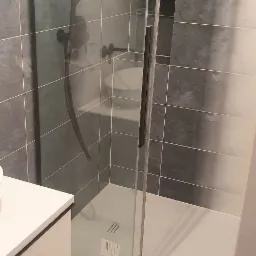 Nowoczesna kabina prysznicowa z czarną armaturą i szarymi płytkami, widoczny fragment białej szafki umywalkowej.