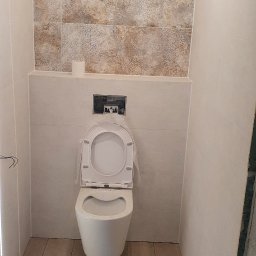 Grzegorz Mazur - Wisząca toaleta z otwartą klapą, zamontowana na tle beżowych płytek z wzorem, z widoczną instalacją i rolką papieru toaletowego, podłoga zabezpieczona kartonem.