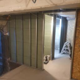 Grzegorz Mazur - Montaż ścian działowych z płyty gipsowo-kartonowej na metalowym stelażu w surowym wnętrzu, widoczna drabina i narzędzia, w tle fragment nieotynkowanej ściany.