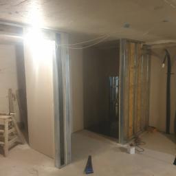 Grzegorz Mazur - Surowy stan poddasza w trakcie adaptacji: widoczne metalowe profile konstrukcyjne, płyty gipsowo-kartonowe, narzędzia i prowizoryczne oświetlenie halogenowe.