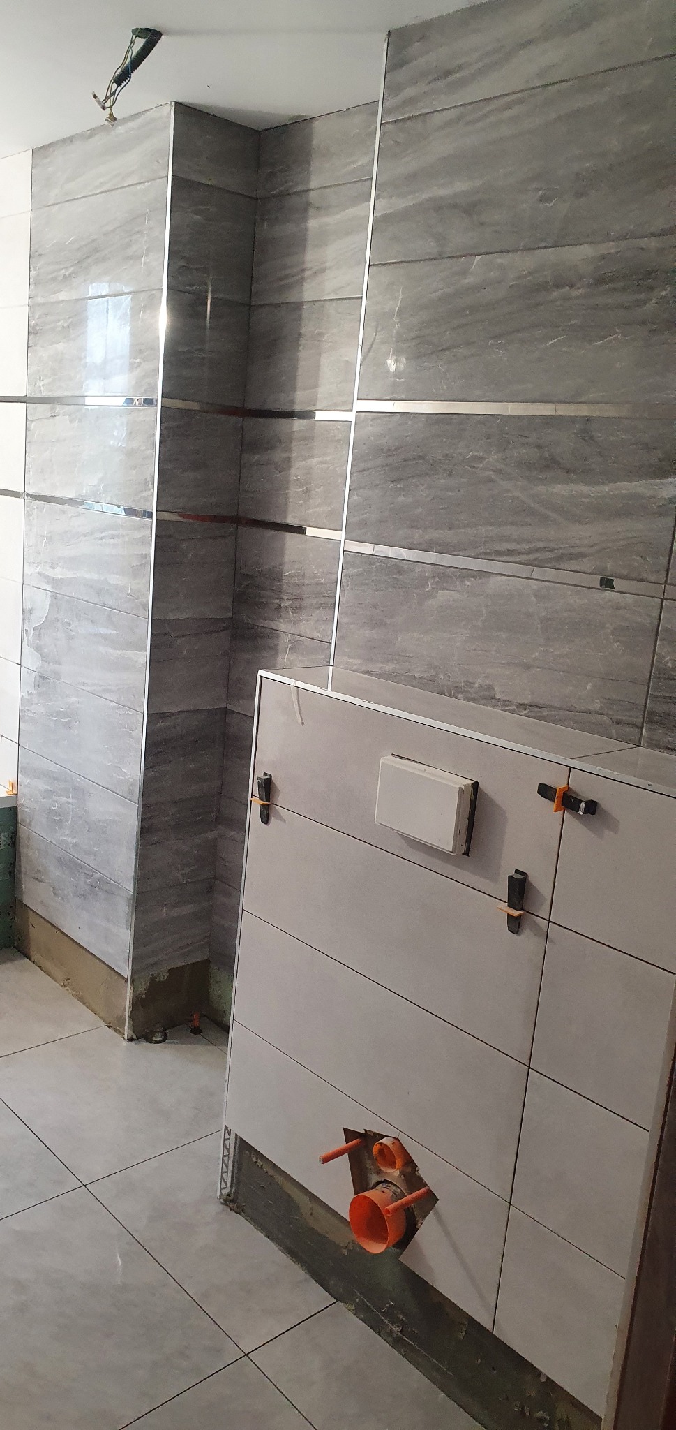 Montaż szarych płytek imitujących marmur na ścianie i pod zabudowę WC z widocznymi elementami instalacji hydraulicznej w łazience.